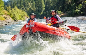 Two people in a bright red raft navigating whitewater rapids, surrounded by lush forest and sunshine.