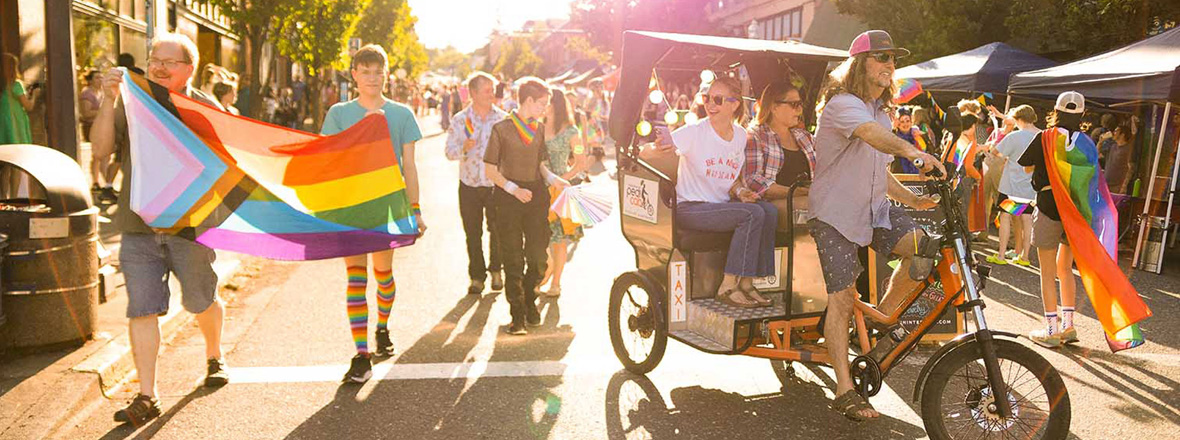 A group of festival goers at a Pride festival. Some hold up a LGBT flag, others ride a taxi bike.