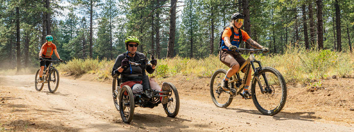 Two cyclists on two-wheeled mountain bikes and one cyclist in a 3-wheeled adaptive bike on a dirt road.