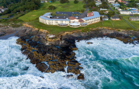 Aerial view of a large oceanfront hotel perched on rocky cliffs along the Oregon Coast with waves crashing below.