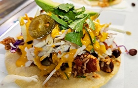 Close-up of a loaded street taco topped with shredded cabbage, rice, beans, jalapeños, cilantro, and creamy orange sauce.