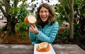 Smiling woman in a teal jacket holding a giant cinnamon roll, seated at an outdoor table.