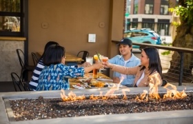 Four friends clinking drinks at an outdoor restaurant patio with a fire feature in the foreground.