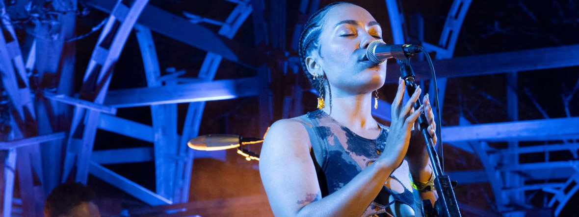 Woman sings into microphone against dark sky and purple lights in background.