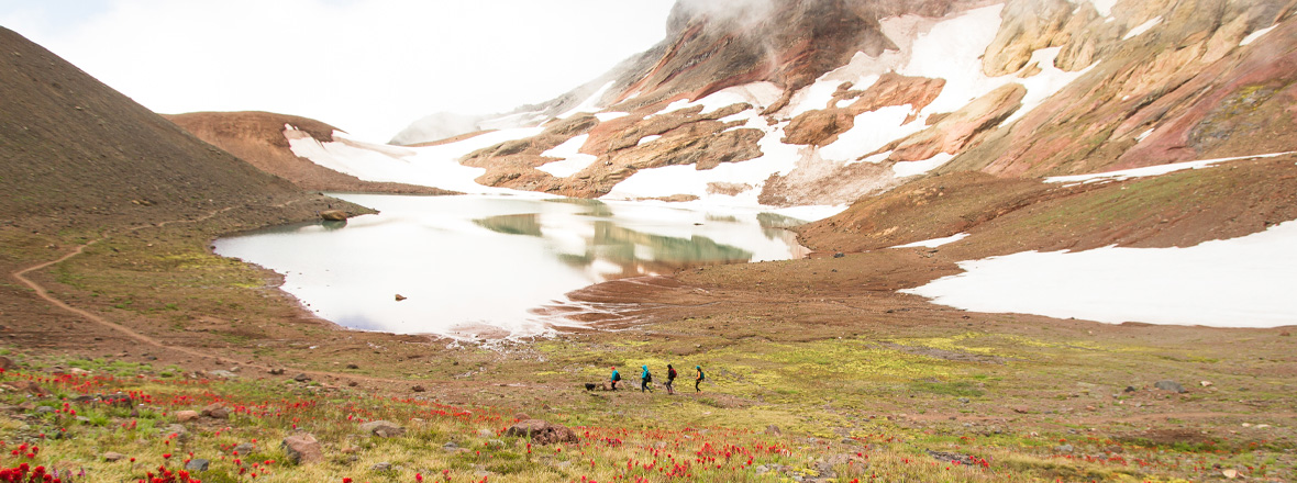 A group of hikers pass a high alpine lake at the base of a snowy mountain.
