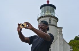 Man smiling as he takes a selfie in front of a historic lighthouse on an overcast day.