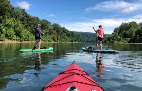 View from a red kayak as two people paddleboard on a calm river surrounded by green trees and blue sky.
