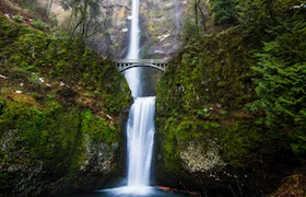 Multnomah Falls cascading in two tiers with a footbridge crossing between the upper and lower falls, surrounded by lush green moss and forest.