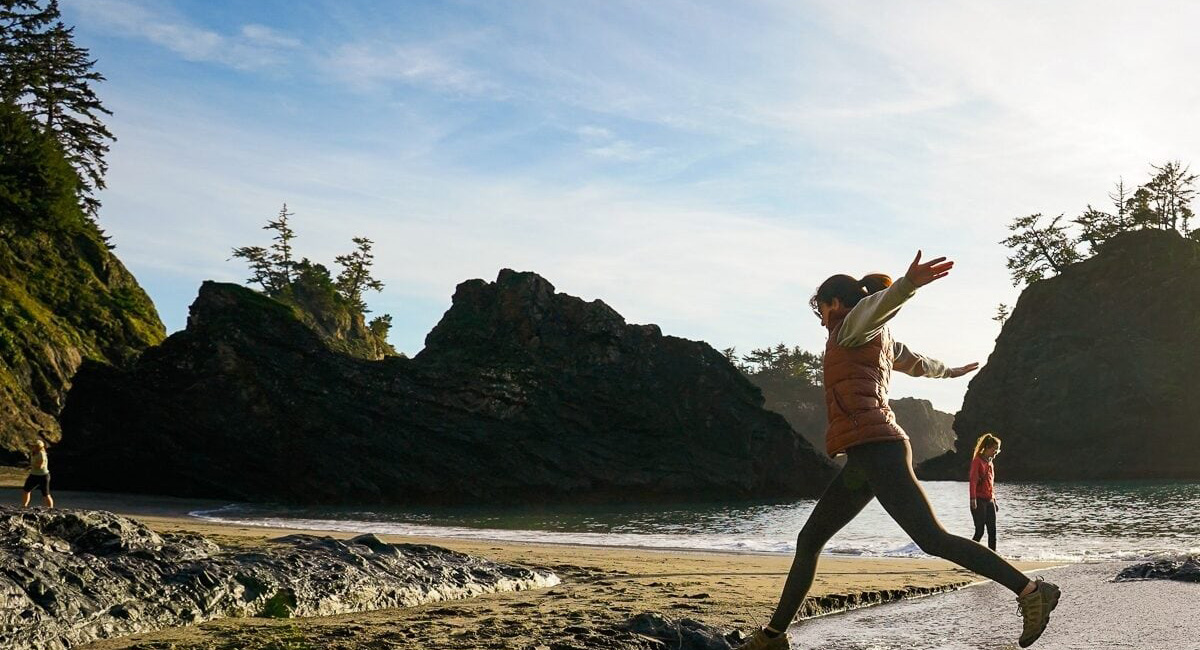 Two people joyfully leaping between rocks on a rugged Oregon Coast beach with sea stacks and forested cliffs in the background.