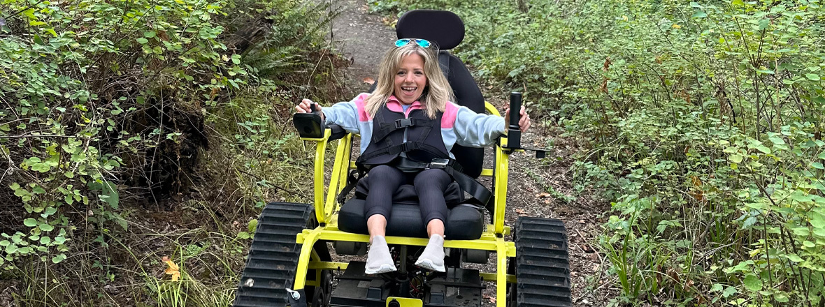 A smiling person navigates a forest trail in a bright yellow all-terrain wheelchair, surrounded by lush green foliage.