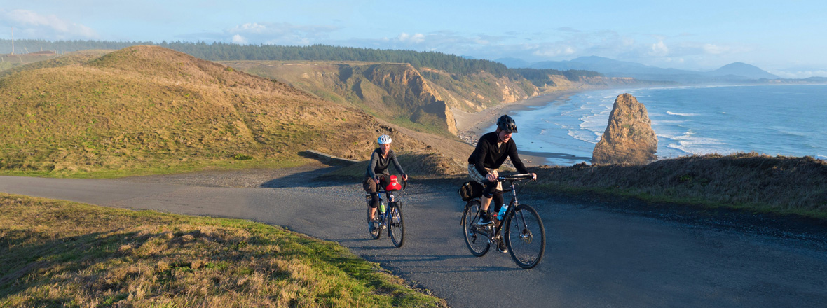 Two cyclists ride up a hill with a scenic coastal backdrop.