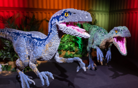 Two colorful dinosaur sculptures made entirely from LEGO bricks, displayed in a dramatic exhibit setting with red and green lighting.
