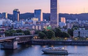 A riverboat cruises along the Willamette River at dusk with downtown Portland in the background, featuring city buildings, a bridge, and soft pastel sky.