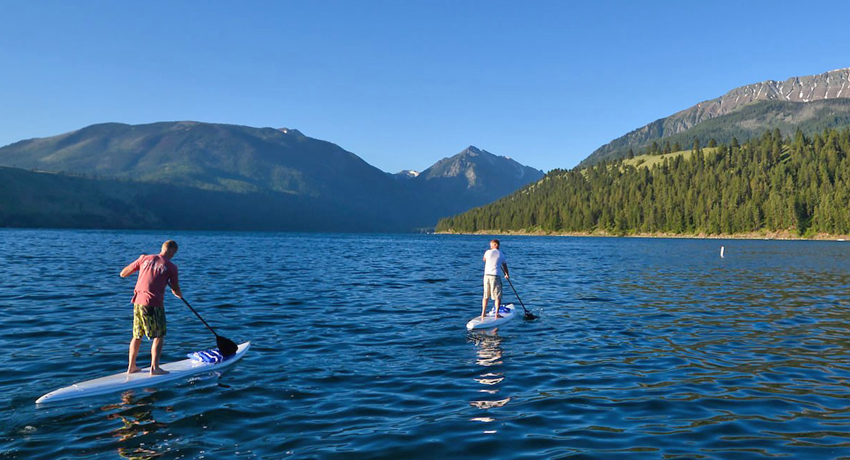 Two people paddleboarding on a calm mountain lake surrounded by evergreen forests and snow-capped peaks under a clear blue sky.