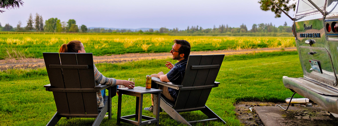 Two people sit on chairs outside of an airstream trailer.