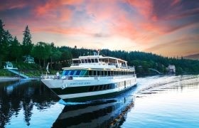A river cruise ship sails through calm waters at sunset, surrounded by forested hills and lakeside homes, with a vibrant pink and orange sky reflecting on the water.