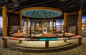 A circular museum exhibit featuring Native American artifacts and interpretive panels, framed by wooden beams and a turquoise ceiling design with Indigenous motifs.