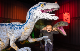A young boy poses playfully with his hands up in front of two large dinosaur sculptures made entirely of LEGO bricks, set in a colorful exhibit.