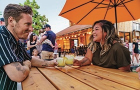 A couple smiles while sharing drinks at a wooden picnic table under a large orange umbrella at an outdoor restaurant.