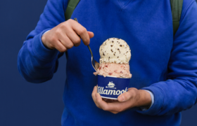 A person in a bright blue sweatshirt holds a cup of Tillamook ice cream with two scoops using a spoon to take a bite.