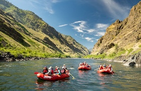 Three red rafts float on a calm river flanked by dramatic canyon walls under a bright blue sky, with paddlers enjoying a group whitewater rafting adventure.