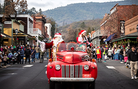 A festive holiday parade with Santa and Mrs. Claus riding a vintage red fire truck down a small-town main street lined with cheering spectators, brick buildings, and forested hills in the background.