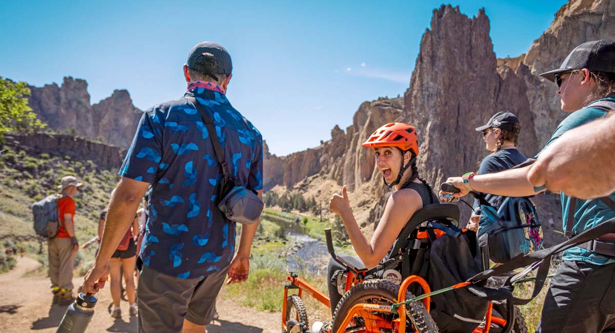 A woman in an adaptive wheelchair gives a thumbs up while on a trail at Smith Rock State Park, surrounded by fellow hikers and dramatic rocky cliffs under a bright blue sky.