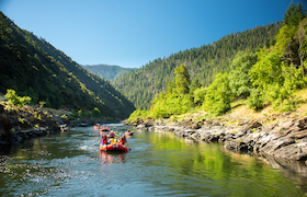 A group of people rafting down a calm section of river surrounded by green forested hills under a bright blue sky.