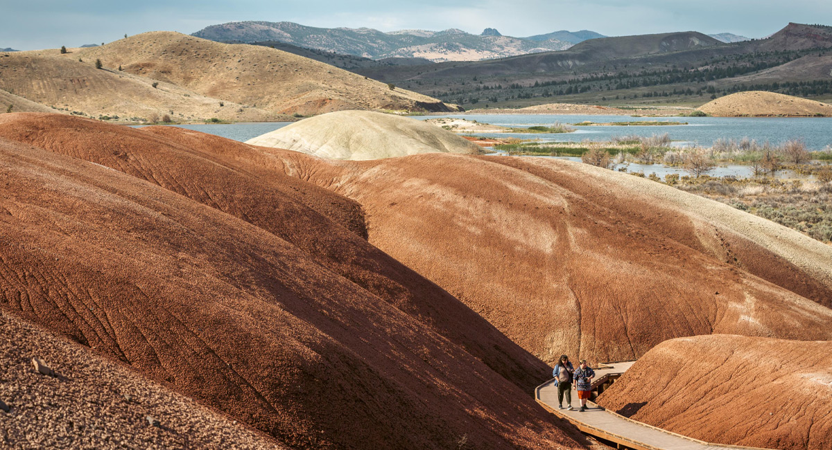 Two people walk on a wood boardwalk through red claystone hills.