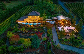 Aerial view of a beautifully lit winery with outdoor seating and lush gardens, surrounded by rows of vineyard at dusk.