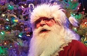Santa Claus smiles in front of a colorful, twinkling holiday tree.