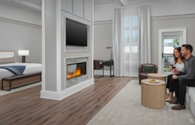 A couple sits by a fireplace in a modern hotel suite, with a bed, flat-screen TV, and large window in view.