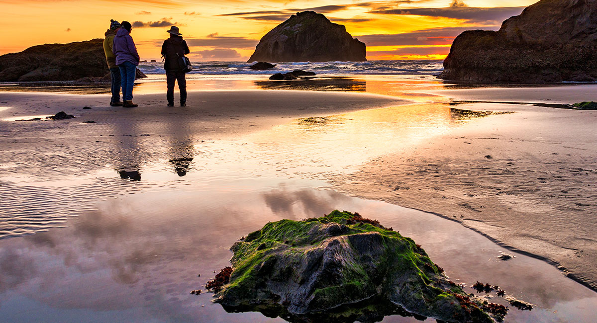 Orange sunset over a sandy and rocky stretch of shore. Two people are visible standing near each other.