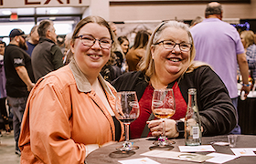 Two women smiling at a table with wine glasses at an indoor event.