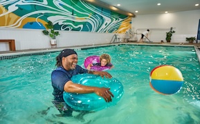Adult and child playing with inner tubes in an indoor swimming pool.