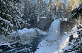Snow-covered forest with a waterfall cascading into a partially frozen river, surrounded by tall evergreen trees.