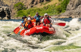 A group of people whitewater rafting in a bright red inflatable boat, paddling through a river surrounded by lush green forest and rocky shoreline.