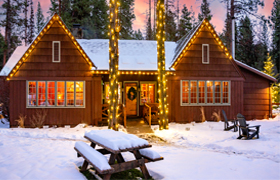 A warmly lit log cabin with snow on the roof and ground, flanked by tall pine trees wrapped in holiday string lights.