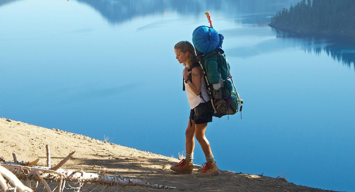 A woman with a large backpack hikes along a steep, dusty trail above a still, blue lake, surrounded by distant forested hills.