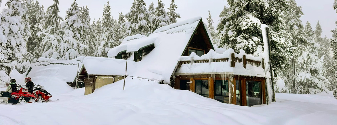 A large wooden cabin covered with snow. A snowmobile is parked near the front entrance.