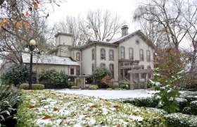 A stately historic house in winter, surrounded by leafless trees and a snow-dusted garden, with patches of green shrubs and leaves peeking through the snow.