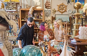 Three people browse shelves filled with vintage nautical collectibles inside an antique store, smiling.