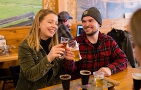 Couple clinking glasses of beer at a wooden table inside a cozy taproom.