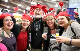 Group of adults wearing red crab hats smile and hold drinks at a lively indoor event.