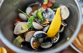 Bowl of steamed clams garnished with a lemon wedge, herbs and sliced vegetables.