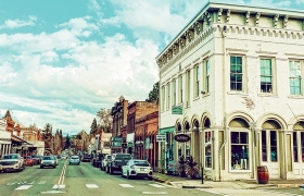 Historic downtown street lined with brick buildings, storefronts and parked cars under a partly cloudy sky.