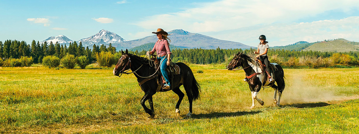 A woman in western wear on a dark brown horse, and a young teen girl on a black and white horse in an open field with views of three mountain peaks.