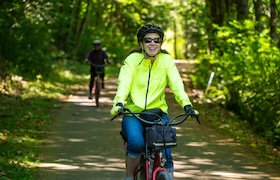 Woman wearing a helmet rides a bicycle along a shaded forest trail with another cyclist behind her.