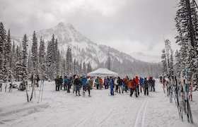 Crowd gathered at a snowy trailhead with skis and snowshoes, with a mountain and pine trees in the background.