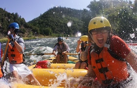 Rafters wearing helmets and life jackets paddle through splashing whitewater on a river.